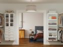 A neatly organized bedroom viewed through an open door, featuring a bed, nightstand, lamp, and adjacent open closet with John Louis Home solid wood storage towers with and shelves full of shoes and clothes.