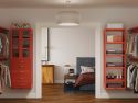 A neatly organized bedroom viewed through an open door, featuring a bed, nightstand, lamp, and adjacent open closet with John Louis Home solid wood storage towers and shelves full of shoes and clothes.