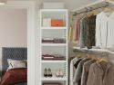 A modern and organized walk-in closet with neatly folded clothes on John Louis Home solid wood shelves, hanging shirts and jackets, a bed with a gray textured headboard and a brown comforter in the background.