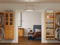 A neatly organized bedroom viewed through an open door, featuring a bed, nightstand, lamp, and adjacent open closet with John Louis Home solid wood storage towers with and shelves full of shoes and clothes.