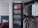 A modern and organized walk-in closet with neatly folded clothes on John Louis Home solid wood shelves, hanging shirts and jackets, a bed with a gray textured headboard and a brown comforter in the background.