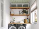 A well-organized laundry room with a washing machine and dryer, John Louis Home solid wooden shelves with decorative plants and storage containers, and a window with a white blind.