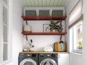 A well-organized laundry room with a washing machine and dryer, John Louis Home solid wooden shelves with decorative plants and storage containers, and a window with a white blind.