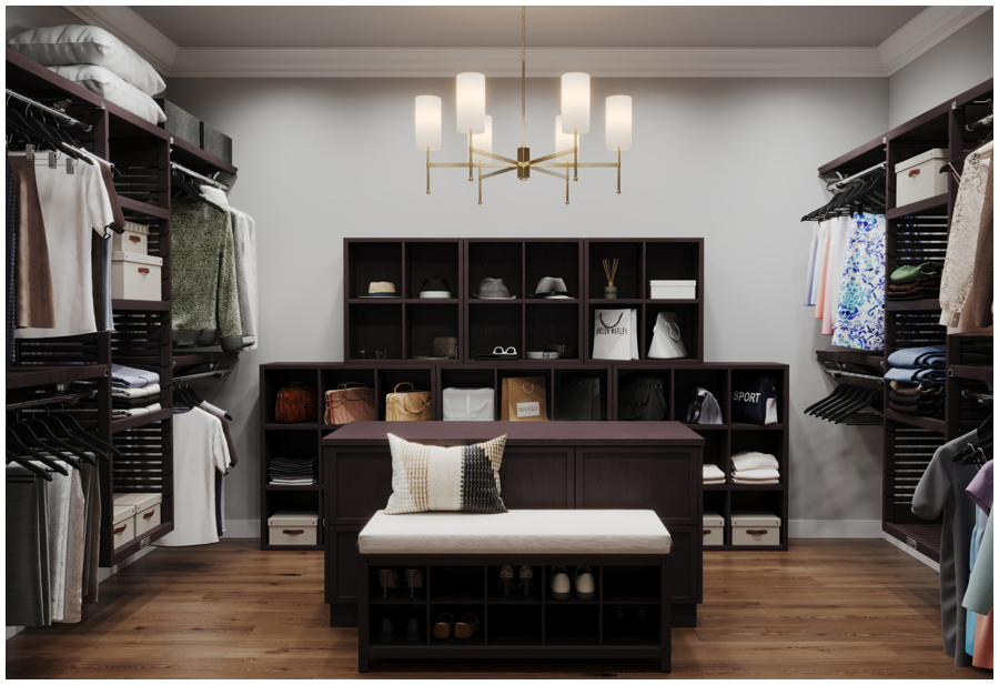 A luxurious walk-in closet with a chandelier hanging from the ceiling. Clothes are hanging on racks and folded on shelves along the walls. There is a bench upholstered in white fabric in the center of the closet.