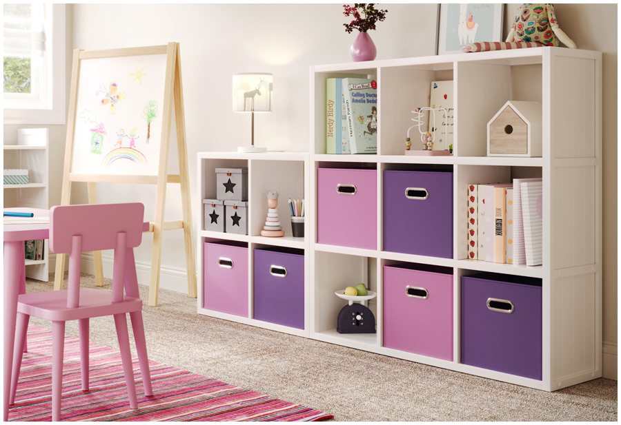 Image of a child’s room interior with a white cube storage against a light colored wall. There are three pink storage bins and two purple storage bins filled with toys and books